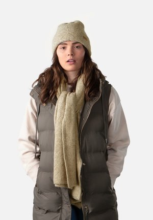 Chaleco acolchado verde sobre un suéter beige, combinado con una bufanda beige texturizada y un gorro de punto beige claro. Materiales suaves e insulated a lo largo.