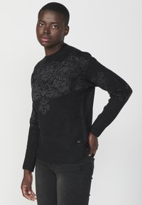 Maglione nero con un design testurizzato e decorato sulla parte anteriore. Presenta un colletto e polsini a coste, realizzato in un morbido materiale lavorato a maglia.