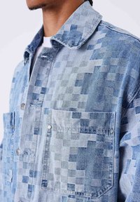 Chaqueta vaquera con un patrón azul pixelado, cuello de camisa, dos bolsillos en el pecho y botones a presión plateados. La textura parece tejida y en capas.
