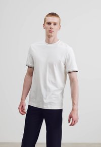 Weißes Baumwoll-T-Shirt mit kurzen Ärmeln, ausgestattet mit schwarzen und marineblauen gestreiften Akzenten an den Bündchen, kombiniert mit marineblauen Hosen.