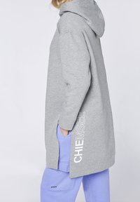 Felpa grigia con cappuccio caratterizzata da una vestibilità comoda, spacchi laterali e un logo stampato in bianco sul lato sinistro. Indossata sopra pantaloni viola.