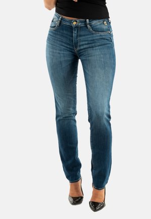 Jeans de mezclilla azul con una cintura de altura media, corte ajustado y un lavado desgastado. Presenta un cierre de botón y bolsillos, combinados con tacones negros.