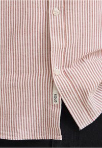 Gros plan sur le devant d'une chemise à rayures verticales rouges et blanches avec deux boutons blancs et une petite étiquette en tissu portant la mention "SOLID" près de l'ourlet.