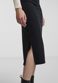 Jupe noire côtelée avec une coupe mi-longue et des fentes sur les côtés. Le matériau semble lisse avec une texture verticale, associé à un style ajusté.