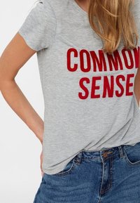 Šedé bavlněné tričko s červeným textem "COMMON SENSE", kulatým výstřihem a krátkými rukávy, spárované s modrými džínovými šortkami.