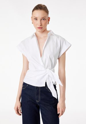 Femme portant un chemisier cache-cœur blanc à manches courtes noué à la taille et un jean en denim bleu foncé, debout devant un fond blanc uni.