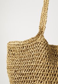Geweven strooien tas in natuurlijke tan kleur, met een gestructureerd oppervlak en een enkele gevlochten schouderband. Rond formaat, ontwerp met open bovenkant.