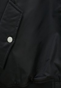 Tissu noir avec un bouton-pression métallique sur une poche à rabat, montrant une texture lisse et légèrement froissée.