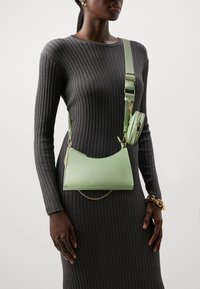 Femme portant une robe côtelée gris foncé avec un sac bandoulière vert clair et une petite pochette assortie, bracelet en chaîne dorée au poignet.