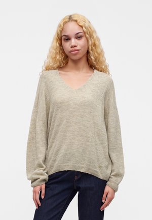 Junge Frau mit langen, lockigen blonden Haaren, trägt einen beigen V-Ausschnitt-Pullover und dunkelblaue Jeans vor einem einfarbigen Hintergrund.