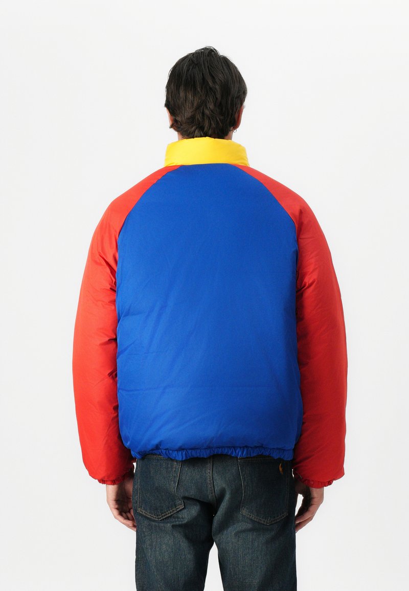 Puchowa kurtka z czerwonymi, niebieskimi i żółtymi panelami, elastycznym dołem oraz wysokim kołnierzem dla ciepła. Materiały wyglądają na gładkie i syntetyczne.