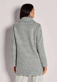 Vrouw met lang, donker haar, gekleed in een middellange, gestructureerde lichtgrijze jas en lichtgekleurde broeken met patroon, met haar rug naar de camera gekeerd tegen een effen achtergrond.