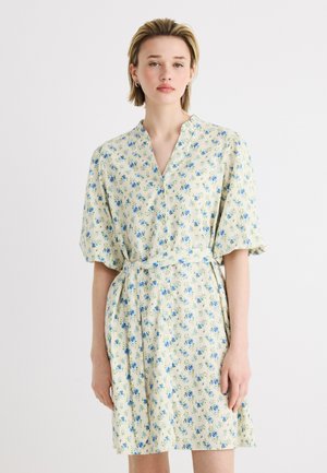 Femme portant une robe courte crème avec un imprimé floral bleu, manches mi-longues, une ceinture nouée à la taille et un col en V.