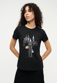 Μαύρο βαμβακερό μπλουζάκι με τυπωμένο σχέδιο μιας αστικής ορίζουσας, μιας γυναίκας με καπέλο και ενός κίτρινου ταξί· κοντά μανίκια και στρογγυλή λαιμόκοψη.