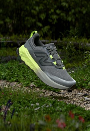 Szürke és neon sárga New Balance futócipő egy sziklás, füves ösvény felett lebeg, zöld növényzet háttérrel.