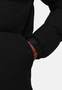 Zwarte pufferjasmanchet met elastische zoom, met een hand in de zijzak, die een gladde textuur en knoopdetail toont.