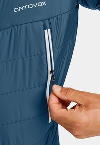 Blaue isolierte Jacke mit vertikalem Steppdesign, kontrastierendem weißen Reißverschluss und praktischer Seitentasche. Hand nahe dem Reißverschluss platziert.