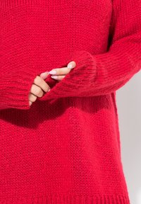 Czerwona dzianinowa sweter z prążkowanymi mankietami, charakteryzująca się luźnym krojem i miękką teksturą. Zbliżenie na ręce częściowo wsunięte w rękawy.