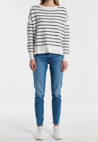 Person in einem weißen Langarmshirt mit schwarzen horizontalen Streifen, blauen Jeans und weißen Sneakern, der vor einem schlichten Hintergrund steht.