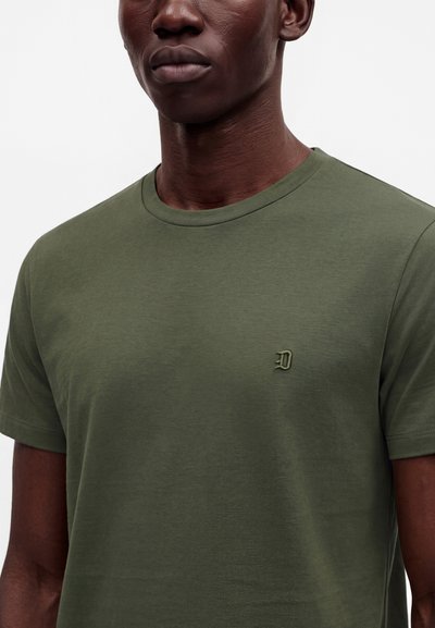 Gros plan d'une personne portant un t-shirt col rond vert olive avec une petite lettre "D" brodée en relief sur le côté gauche de la poitrine.