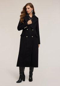 Cappotto doppiopetto nero con dettagli in bottoni dorati, ampi rever e due tasche frontali, abbinato a stivali neri lucidi al ginocchio.