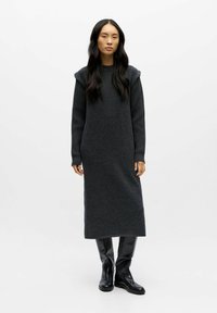 Abito lungo in maglia grigia scuro con maniche a coste, caratterizzato da una vestibilità comoda e dettagli sulle spalle. Abbinato a stivali neri al ginocchio.