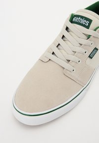 Beige suede sneaker with green and white accents, featuring flat laces, a padded tongue, and a textured rubber sole. Etnies branding visible.