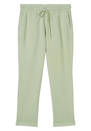 Persoon die lichtgroene joggingbroek met trekkoord, olijfgroene trui en witte sneakers draagt, staand tegen een witte muur.