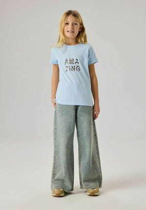 Enfant portant un t-shirt graphique bleu clair avec l'inscription "Amazing", un jean gris à jambes larges, et des baskets beige, debout devant un fond neutre.