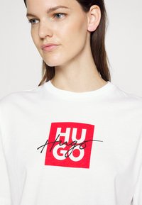 Biela bavlnená tričko s odvážnym červeným obdĺžnikovým logom, na ktorom je v bielej farbe napísané "HUGO" a v čiernom kurzíve "Hugs". Okrúhly výstrih, krátke rukávy.