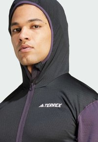 Giacca con cappuccio nera e viola, caratterizzata da una chiusura con zip, cappuccio regolabile e logo "TERREX" sul petto, realizzata in un tessuto testurizzato.