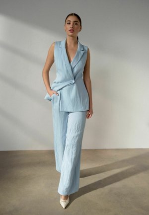 Sieviete stāv pret vienkāršu sienu, tērpusies gaiši zilā bezroku žaketē un atbilstošās plaša piegriezuma biksēs, valkājot baltas smailas kurpes uz papēža.