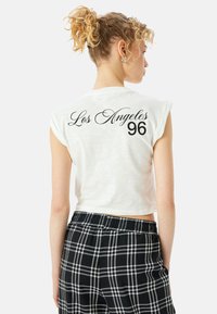 Camiseta blanca recortada con mangas enrolladas que presenta el texto negro "Los Ángeles 96" en la parte posterior. Combinada con pantalones de cuadros en blanco y negro.