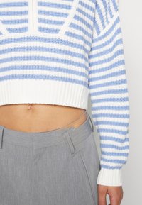 Addome scoperto con un maglione corto a righe blu e bianche, pantaloni grigi a vita alta e una catena argento in vita.