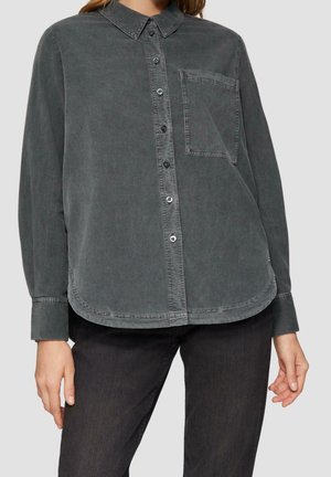 Woman wearing a faded black button-up shirt with a chest pocket, paired with dark jeans, standing against a plain light gray background.