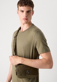 Olíva zöld keresztpántos táska letisztult dizájnnal, fekete kiegészítőkkel és textúrált pánttal, egy modelltól, aki egy megfelelő T-shirt-ot visel.
