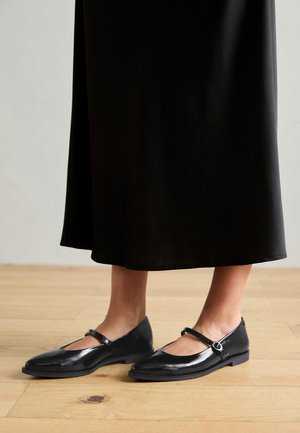 Černé lakované baleríny s páskem Mary Jane, špičatou špičkou a jemným leskem, doplněné o splývavou černou sukni na dřevěné podlaze.