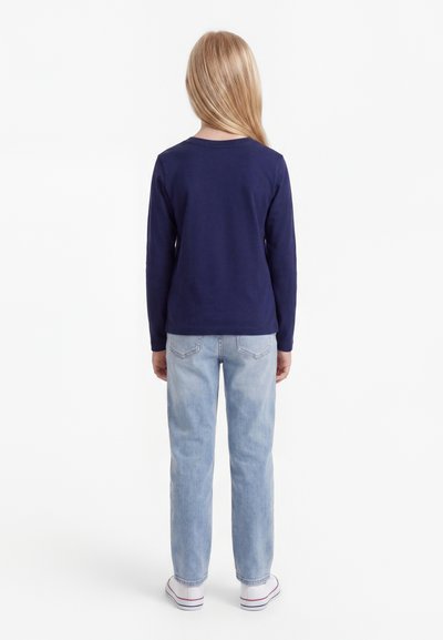 Dieťa s dlhými blond vlasmi, oblečené v námorníckej modrej tričku s dlhým rukávom, svetlomodrých džínsach a bielych teniskách, stojace chrbtom na bielom pozadí.