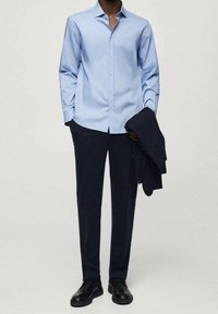 Uomo in camicia azzurro chiaro, pantaloni blu scuro, che tiene in mano un blazer coordinato scuro e scarpe eleganti nere, in piedi su uno sfondo semplice.