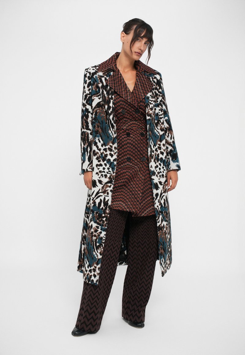 Lange jas met een zwart, wit en blauw bloemmotief; maroon zigzag dubbelgebreid jurkje eronder; brede zigzag broek. Glad textuur.