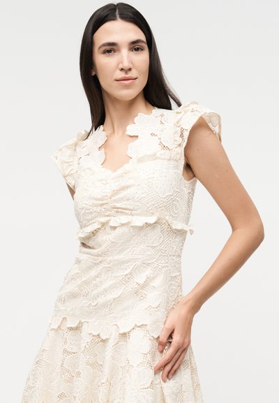 Γυναίκα με μακριά σκούρα μαλλιά που φοράει ένα κρεμ, λουλουδάτο, δαντελωτό φόρεμα με κοντά μανίκια με βολάν, στέκεται μπροστά σε απλό φόντο.
