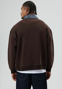 Bruine sweatshirt met een denim kraag, met een ontspannen pasvorm en geribbelde boorden. De achtergrond is effen voor een minimalistische uitstraling.