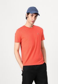 Tricou scurt, de culoare coral strălucitor, din bumbac, cu guler rotund și un accent cu un logo mic. Este purtat cu pantaloni negri și o pălărie albastră.