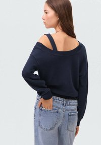 Een model met lang bruin haar draagt een navy off-shoulder trui en lichtblauwe jeans, staand met de handen in de zakken, lichtjes van zich af gekeerd.