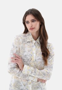 Camicia floreale a maniche lunghe in tessuto bianco trasparente con stampa botanica gialla e grigia, dotata di colletto a punta e polsini con bottoni.