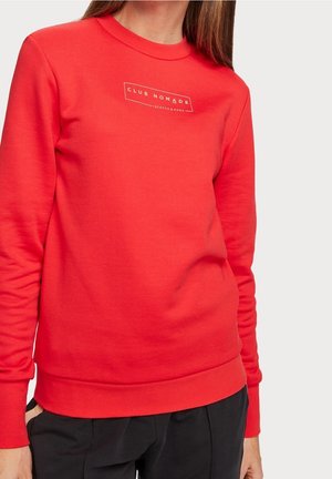 Röd sweatshirt i mjukt tyg; har en rund halsringning och en rektangulär vit "CLUB NOMADE"-logotyp på bröstet.