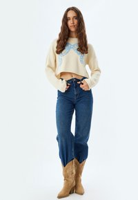 Pullover corto in maglia crema con design a fiocco blu, abbinato a jeans blu a vita alta e stivali da cowboy beige con cuciture decorative.