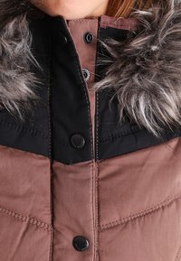 Brown puffy jacket with black accents, featuring a fur-lined collar, snap buttons, and quilted texture. Close-up view of front.