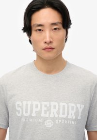 Grå bomuld t-shirt med stort hvidt "SUPERDRY" logo-tekst, klassisk rund halsudskæring og korte ærmer. Simpelt, afslappet design.