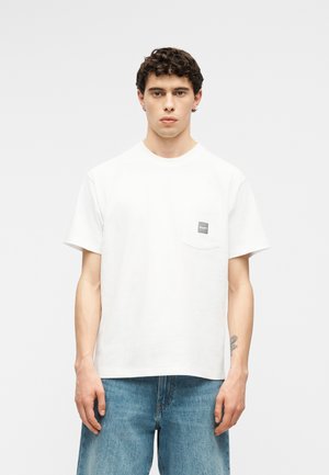 Noor mees lühikeste lokkis juustega, kes kannab tavalist valget T-särki väikese rinnataskuga ja siniseid teksapükse valge tausta ees.